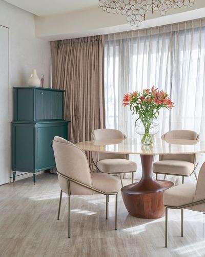 Oval marble dining table with sculpted wood base, cane chairs, and a green cabinet by sheer curtained window
