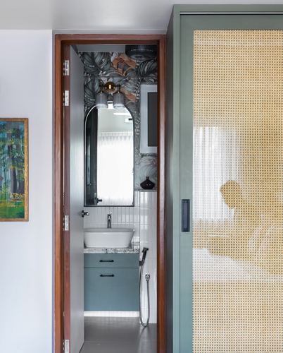 View through wood-framed doorway into bathroom with arched mirror, green cabinetry, and botanical wallpaper, cane door beside