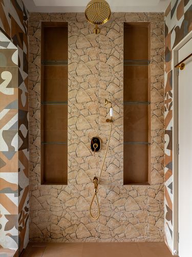 Shower wall in crazy-cut stone with brass fittings, flanked by niches with glass shelves.