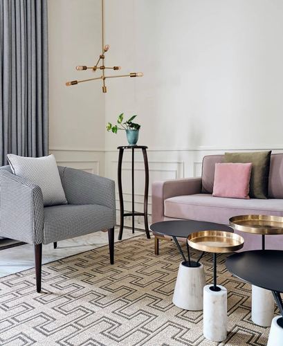 Living room corner with mauve velvet sofa, houndstooth armchair, brass chandelier, and marble-base tray tables on geometric rug