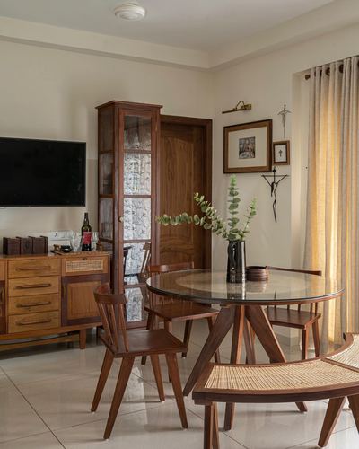 Dining area with round glass table, wooden chairs, cane bench and antique cabinet against cream walls