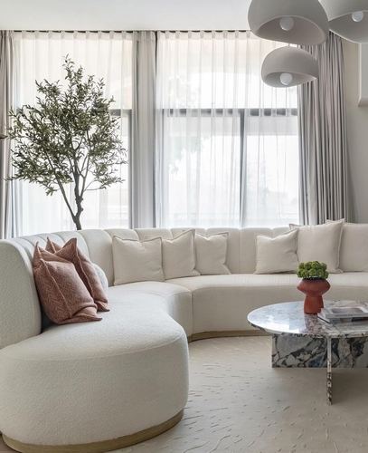 Curved cream boucle sectional sofa with marble coffee table and olive tree by sheer-curtained window