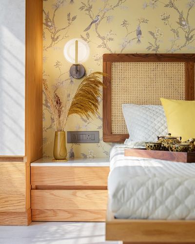 Bedroom corner with cane headboard, bird-patterned wallpaper, and a wooden nightstand with dried pampas grass