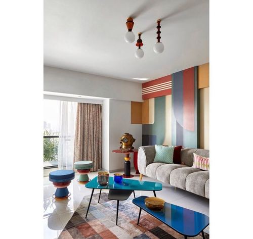 Colorful Memphis-style living room with blue glass tables, grey sofa, and geometric mural wall