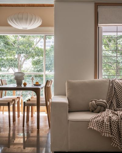 Beige sofa beside dining area with wooden table and cocoon pendant light, large windows showing trees