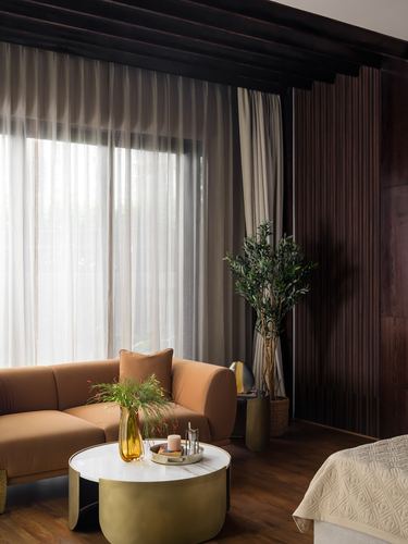 Terracotta sofa and brass round coffee table beside sheer-curtained window in wood-paneled living room