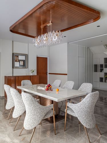 Dining room with marble table, boucle chairs, and chandelier under a wood ceiling recess