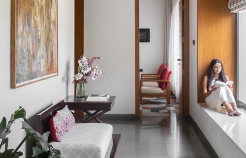 Woman reads on a window seat at the end of a sunlit corridor with wooden doors and a painted living room beyond