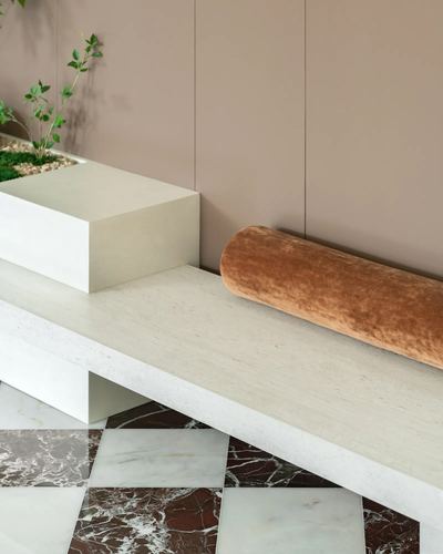 Travertine bench with velvet bolster, planter, and burgundy-white marble checkered floor against blush wall