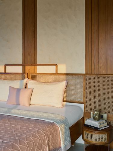 Bed with cane headboard, pink cushions, and round cane nightstand against textured plaster and wood wall