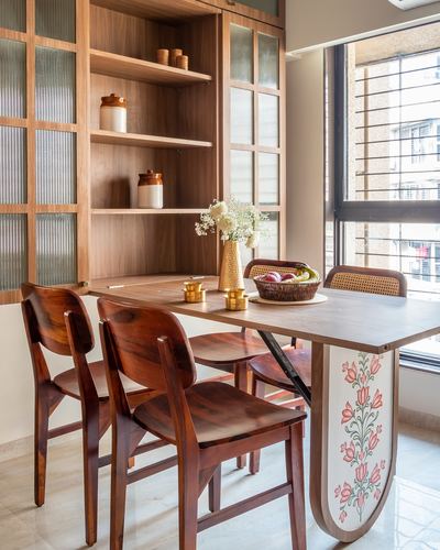 Wooden dining table with cane chairs beside a floral-painted panel and glass-front cabinetry