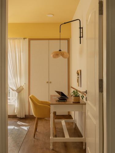 Doorway view of a small yellow study nook with wardrobe, desk, mustard chair and woven pendant light