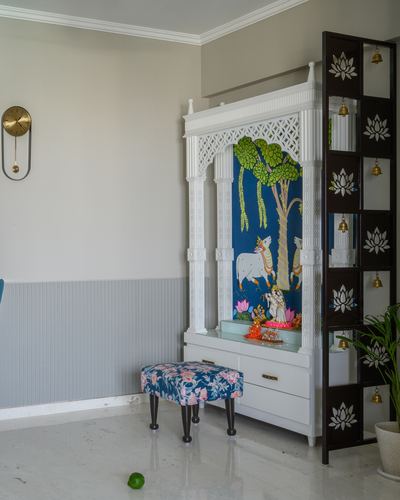 White carved pooja unit with blue Krishna mural, lotus bell screen, and floral stool on marble floor