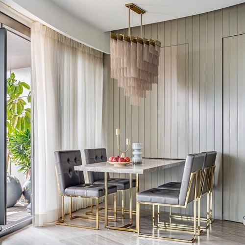 Dining nook with marble table, gold-framed grey chairs, and fringe chandelier against slatted wall