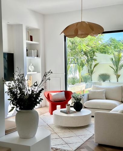 White living room with red curved chair, round stone coffee table, and petal-shaped pendant light near a garden view