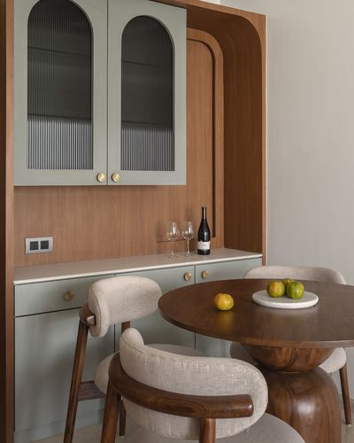 Sage-green arched glass cabinet above wood cabinetry beside round wooden dining table with upholstered chairs