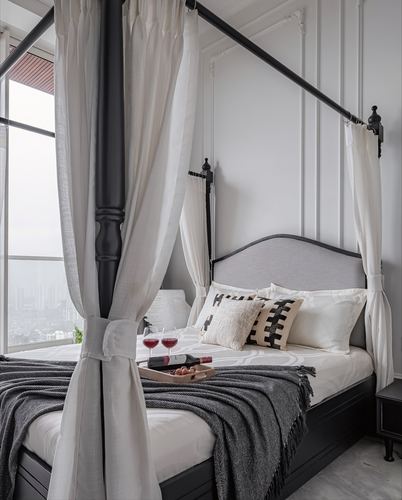 Four-poster bed with sheer curtains, gray headboard, and wine tray, city view through window