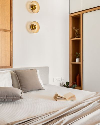 Bed with taupe cushions and open book beside wood shelving and brass wall sconces