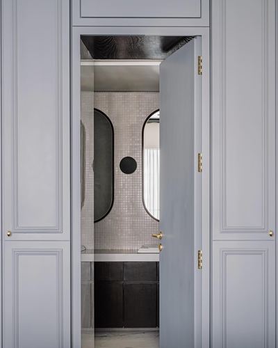 Grey paneled door ajar revealing bathroom with twin oval mirrors and mosaic tile wall
