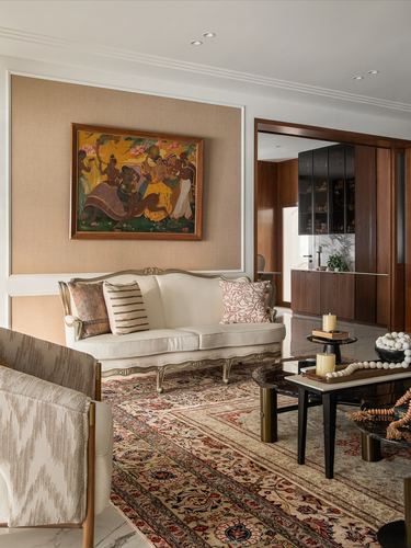 Living room with carved cream settee under a framed painting on an upholstered panel, patterned rug, dark coffee table.
