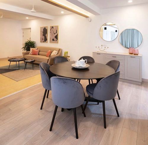 Round dark wood dining table with grey velvet chairs, sideboard and circular mirrors behind, living area beyond