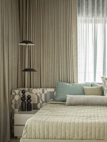 Bedroom corner with pleated linen curtains, dark pendant lamps, patterned bench and quilted bed.