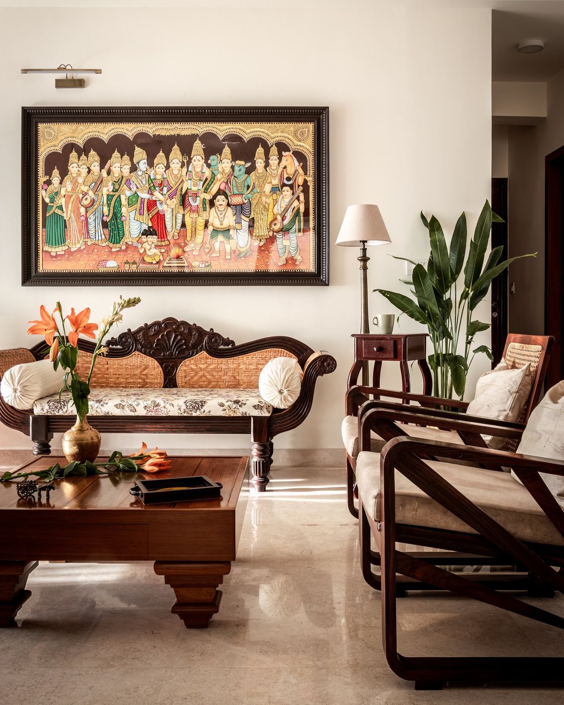 Living room with carved wooden cane sofa, coffee table, framed traditional painting, plant, and lamp on marble floor