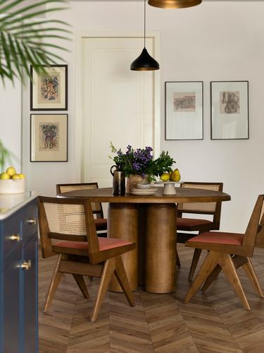 Round wood dining table with cane chairs beneath a black pendant, framed art on walls