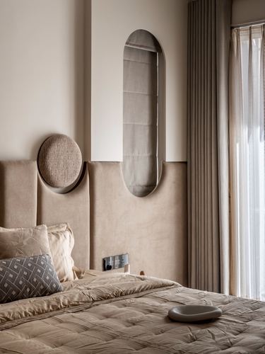 Beige upholstered headboard with oval mirror and round sconce beside curtained window