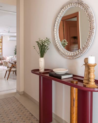 Maroon console table below a round carved mirror in a foyer, styled with vase, bowl and candle holders