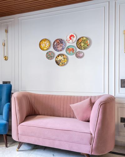Blush pink channel-tufted sofa below a cluster of decorative wall plates, blue chair beside it