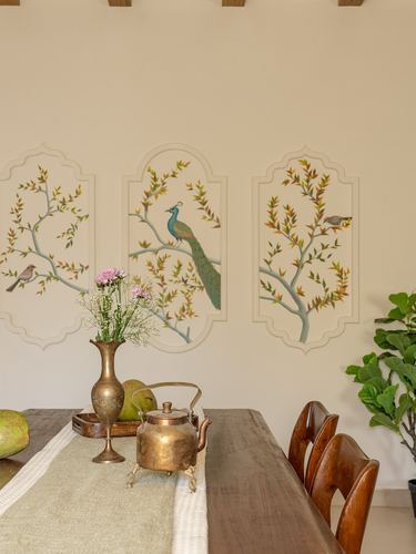 Dining table with brass vase and kettle beneath hand-painted peacock wall panels