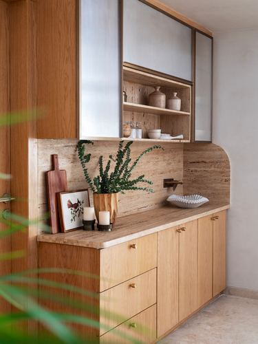 Wood cabinetry kitchen corner with travertine backsplash, brass fixtures, plant and ceramic decor