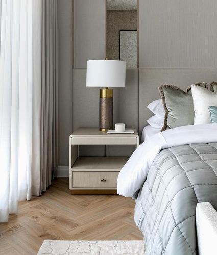 Bedroom corner with upholstered grey panelling, nightstand, brass table lamp, and quilted bedding on herringbone floor
