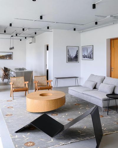 Open-plan living room with round wood coffee table, cane chairs, and exposed ceiling conduits in Mumbai apartment