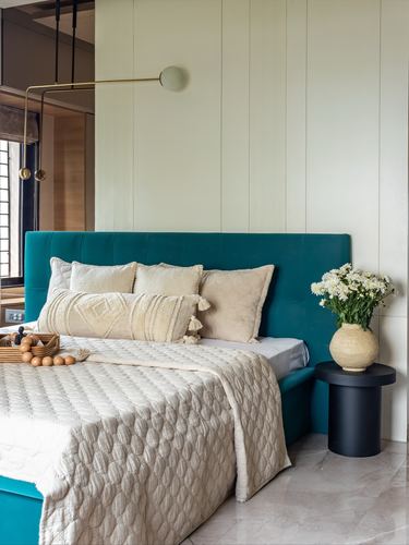 Bedroom with teal tufted headboard, cream bedding, brass sconce, and round black side table with vase