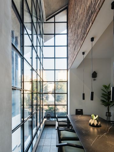Double-height steel-framed glass wall beside dark stone dining table with pendant lights