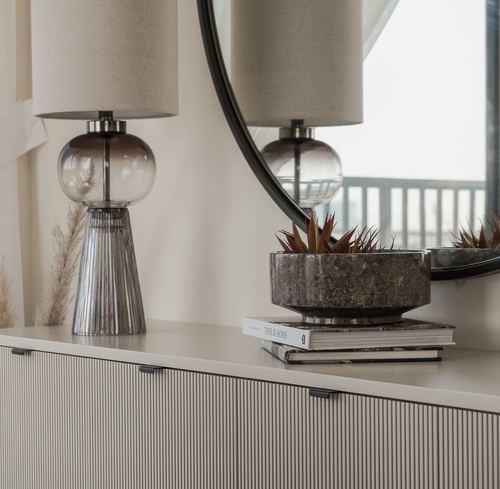 Sideboard vignette with glass table lamp, round mirror, and stone planter on stacked books