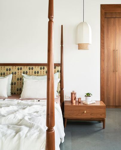 Wooden four-poster bed beside cane-shaded pendant light and wood nightstand in a warm-toned bedroom.
