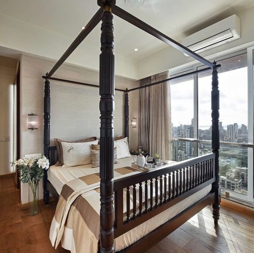 Dark wood four-poster bed with railing footboard facing floor-to-ceiling windows overlooking a city skyline