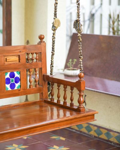 Close-up of a carved wooden jhula with brass chains and a painted tile inlay on a sunlit porch