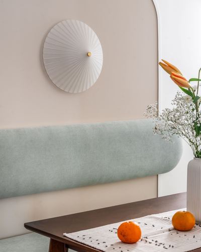Sage green curved banquette below a pleated fan wall light, with flowers and oranges on table