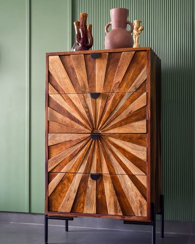 Wooden chest of drawers with sunburst marquetry pattern against a green ribbed wall, topped with ceramic vases