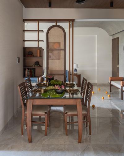 Wooden dining table set with brass bowls beside an arched cane-screen partition in Ahmedabad home