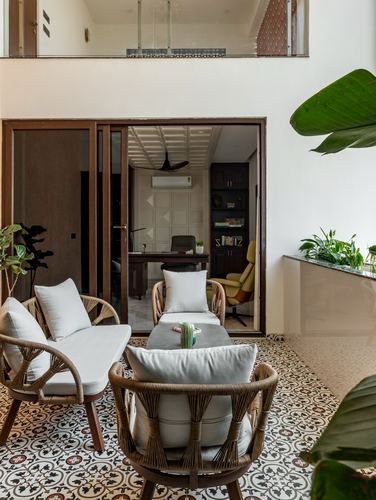 Cane armchairs on patterned tile floor in a double-height courtyard seating area beside a glass-walled study