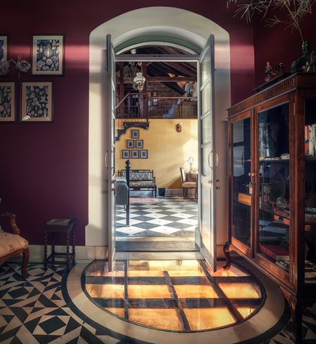 View through an open arched doorway from a maroon room into a sunlit hall with checkerboard floor in a heritage Goan house