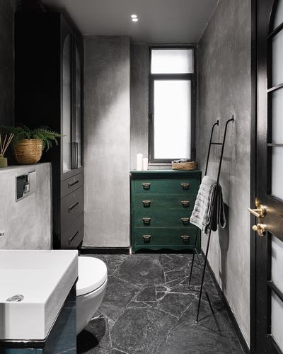 Dark bathroom with textured grey walls, green vintage dresser, stone floor and black metal towel ladder