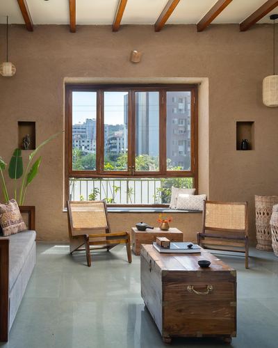Living room with exposed wooden rafters, plastered walls, cane chairs and a wooden trunk coffee table facing a window with city view.