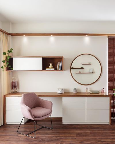 Study nook with wooden desk, pink chair, and round mirror with shelves against a cream wall.