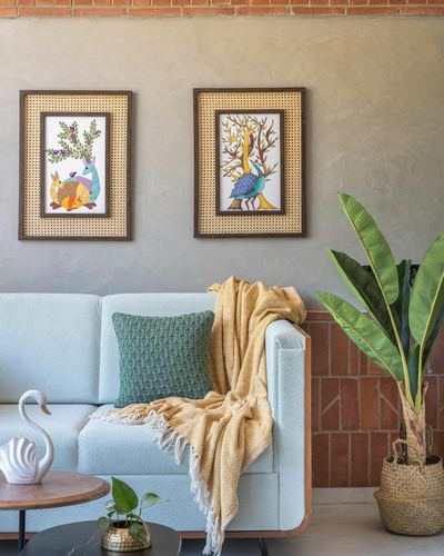 Pale blue boucle sofa with mustard throw beneath two cane-framed gond art paintings on plastered wall
